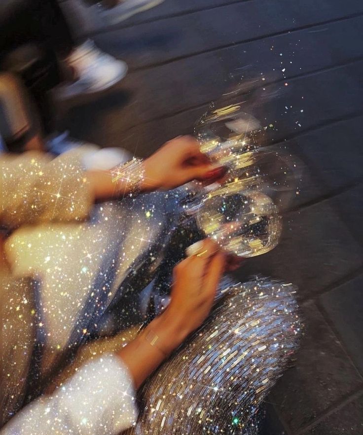 Person in a sparkling outfit holding two sparkly wine glasses on a dark floor.