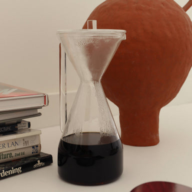 Hourglass Carafe with dark liquid next to a terracotta vase on a white surface