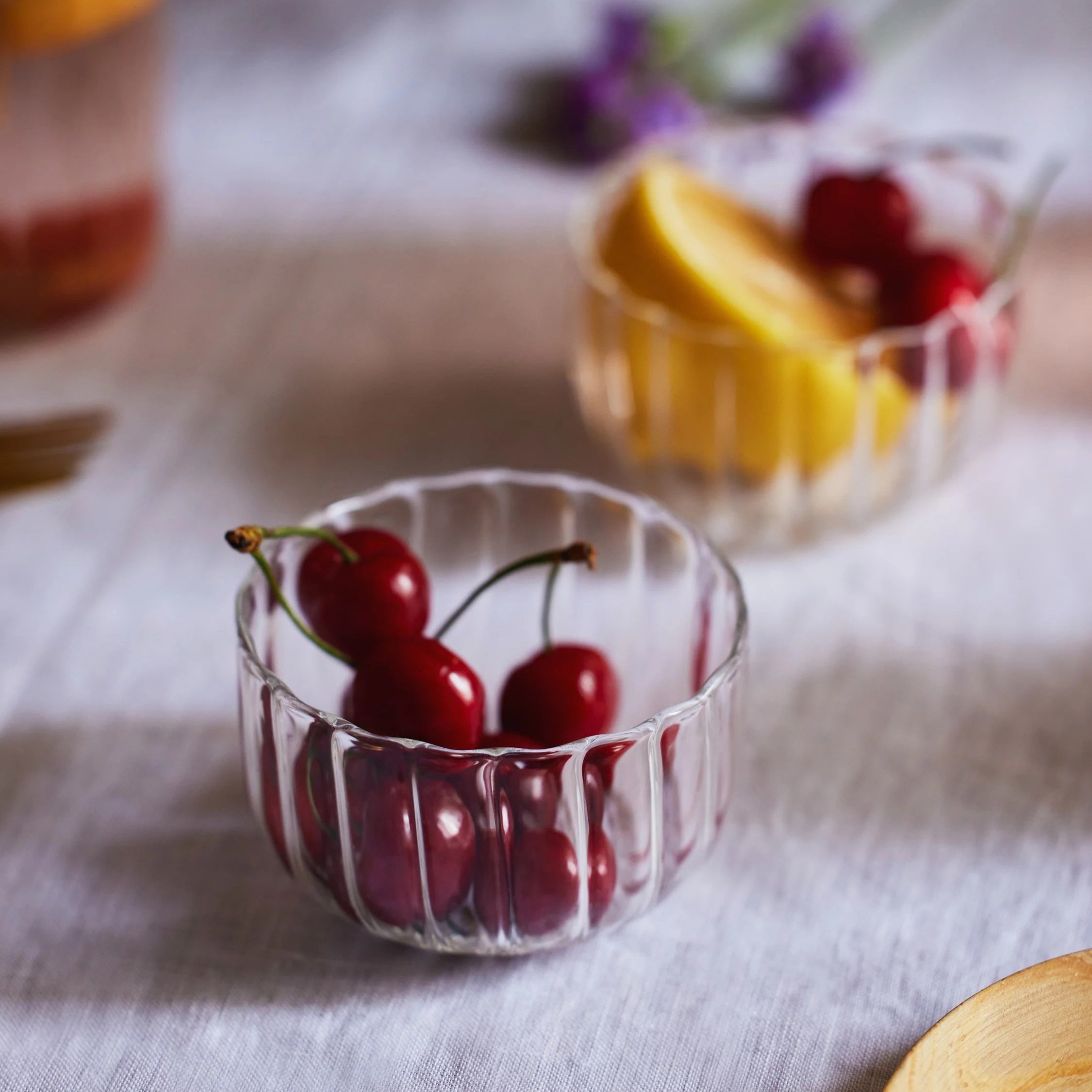Clear glass bowls with cherries and fruit on a light fabric surface.